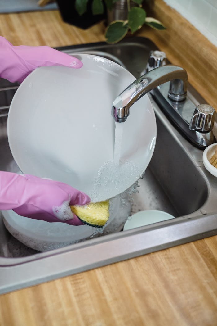 Hands in pink gloves washing dishes in a stainless steel kitchen sink under running water.